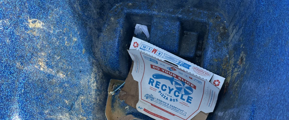 An empty Domino's pizza box with a 'Recycle This Pizza Box' stamp, inside a textured blue recycling bin.