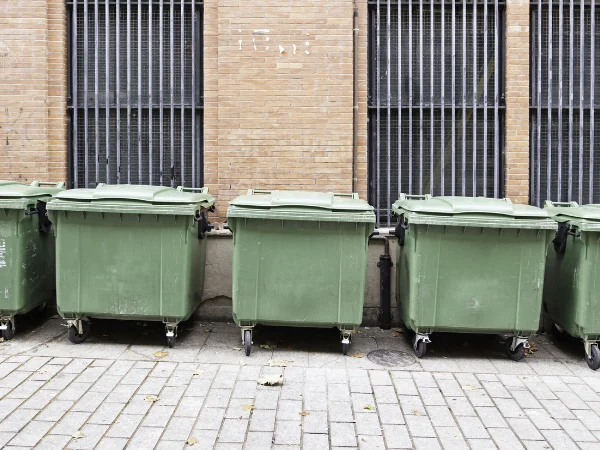 Several large green commercial dumpsters line a brick wall with barred windows on a paved sidewalk.
