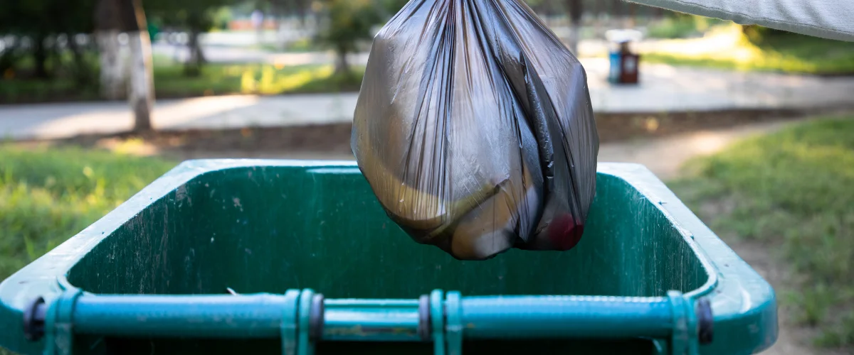 A black trash bag is being lowered into a green outdoor trash bin on a sunny day with green grass in the background. A black trash bag is being lowered into a green outdoor trash bin on a sunny day with green grass in the background.