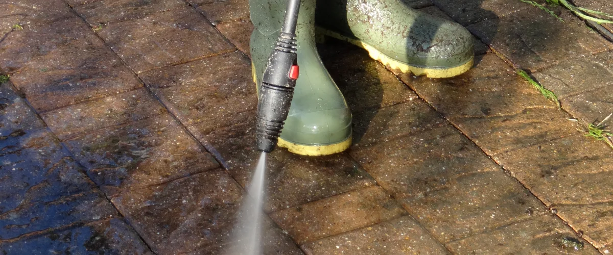 A person in green wellington boots using a power washer to clean dirty, brown patio pavers. A person in green wellington boots using a power washer to clean dirty, brown patio pavers.