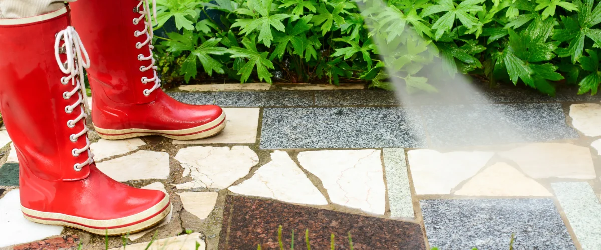 A person in shiny red rain boots with white laces stands on a stone path next to green plants being watered. A person in shiny red rain boots with white laces stands on a stone path next to green plants being watered.