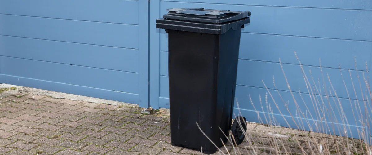 A black plastic waste bin with wheels stands upright on a brick patio next to a blue horizontal slat gate.