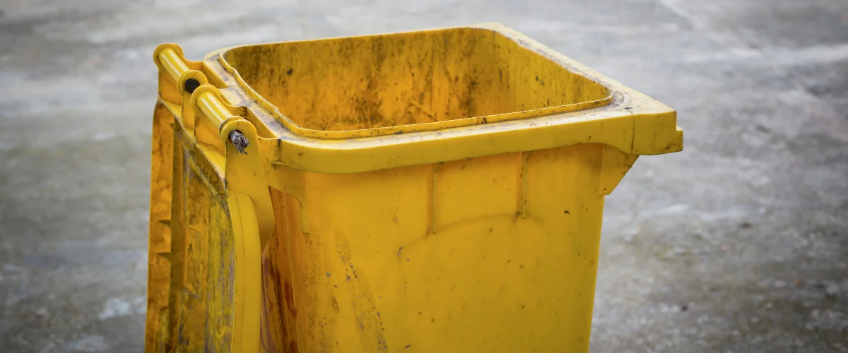A weathered, grimy yellow trash bin with an open lid, sitting on a blurred concrete surface.