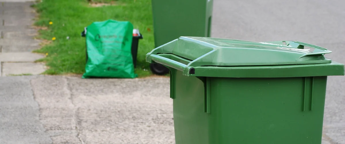 A close-up of a green recycling bin lid with a green bag and another bin in the blurry background on a sidewalk. A close-up of a green recycling bin lid with a green bag and another bin in the blurry background on a sidewalk.