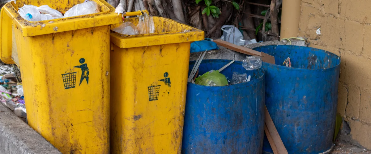 Two overflowing yellow plastic bins and two blue barrels, all full of trash, sit outdoors next to a concrete wall.