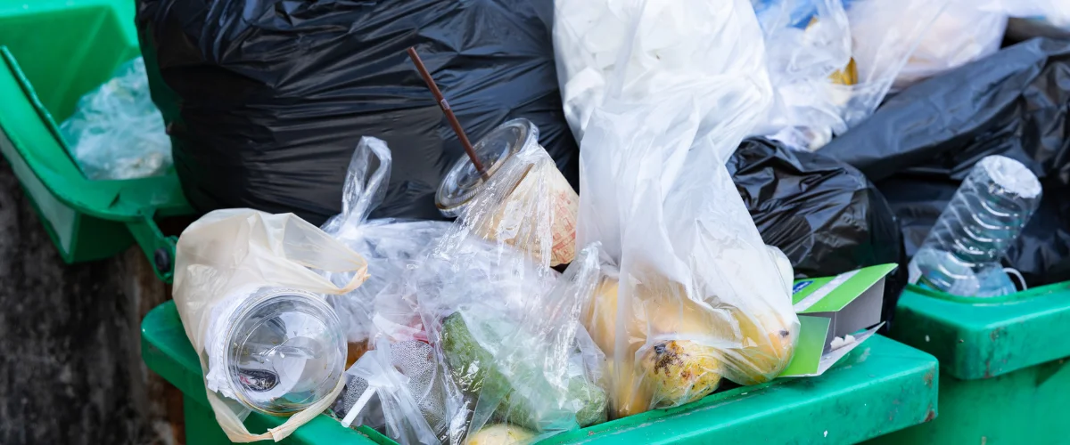 A green dumpster overflowing with plastic bags, bottles, and food waste, highlighting excessive plastic packaging.