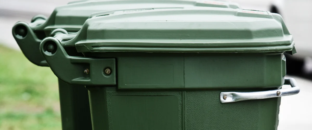 Close-up of two dark green plastic garbage bins with hinged lids and a metal handle on the right bin.