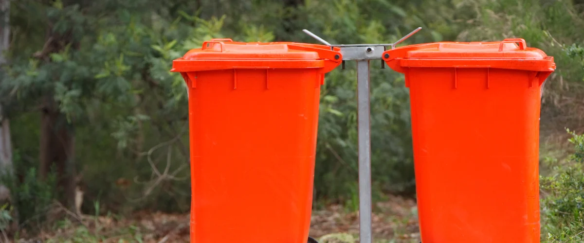 Two bright orange garbage bins are connected by a metal frame against a blurred background of green trees and foliage.