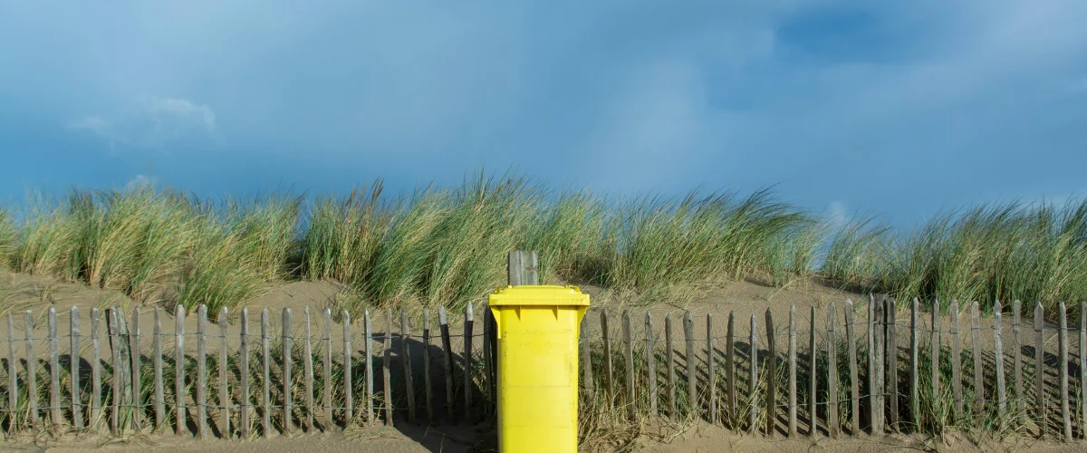 A bright yellow recycling bin stands in front of a wooden fence, sand dunes with green grass, and a blue sky. A bright yellow recycling bin stands in front of a wooden fence, sand dunes with green grass, and a blue sky.
