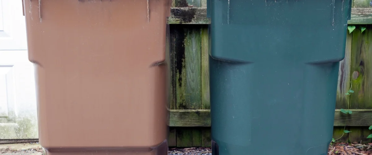 A brown and a dark green garbage can standing side-by-side against a weathered wooden fence. A brown and a dark green garbage can standing side-by-side against a weathered wooden fence.