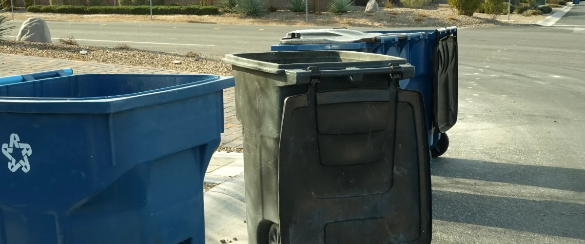 A row of blue and black trash and recycling bins lined up next to a paved street on a sunny day. A row of blue and black trash and recycling bins lined up next to a paved street on a sunny day.