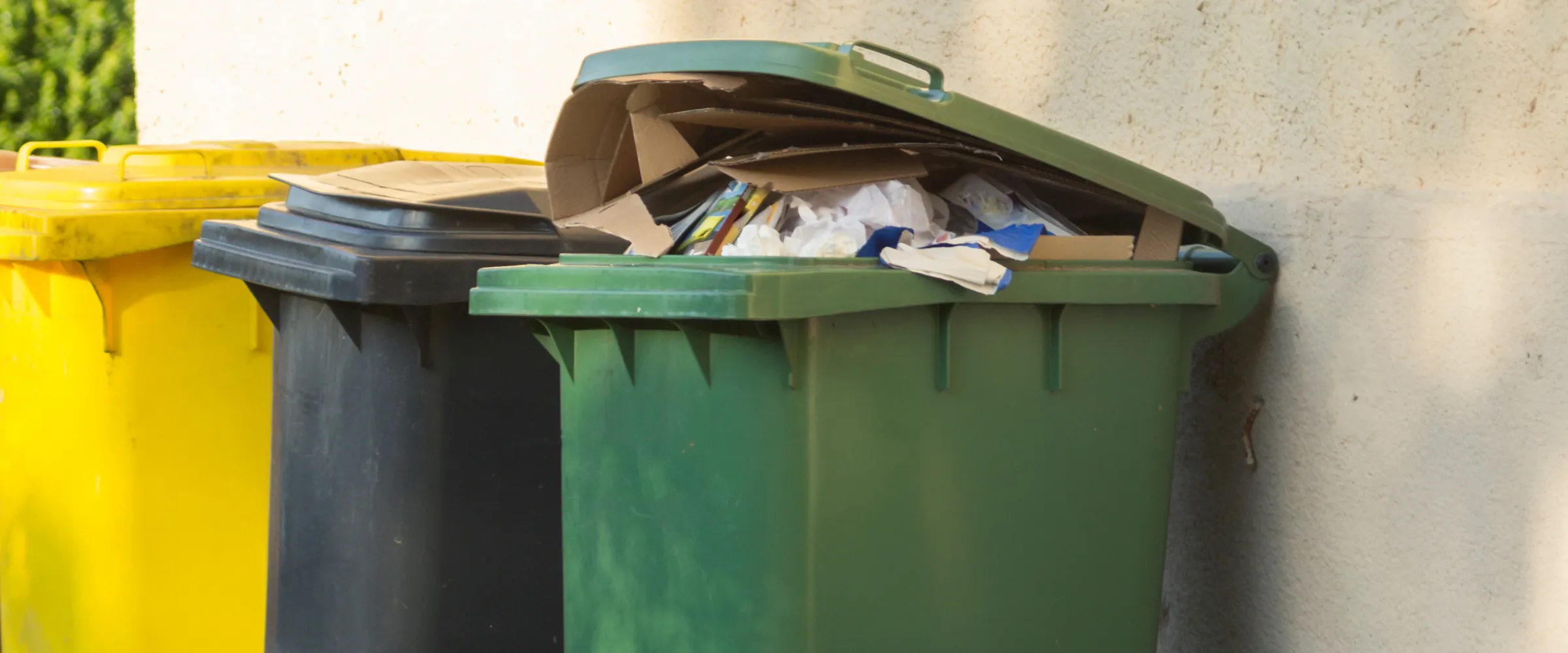 An overflowing green recycling bin with its lid open, full of cardboard and plastic, next to a black and yellow bin. An overflowing green recycling bin with its lid open, full of cardboard and plastic, next to a black and yellow bin.