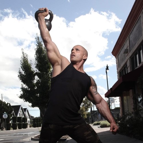 A muscular bald man lifting a kettlebell overhead outdoors on a city street, wearing a black tank top and camo pants.
