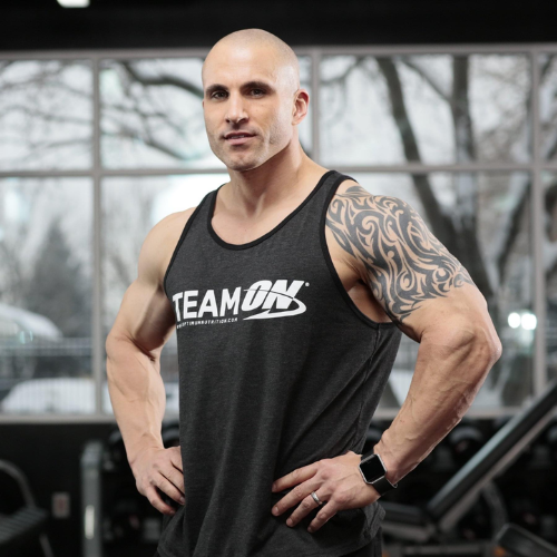 Portrait of a muscular bald man wearing a black tank top with 'TEAM ON' logo, holding a kettlebell in a dimly lit gym with tires and a brick wall.