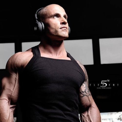 Photo of a bald man with an athletic build, wearing a grey t-shirt with 'TEAM ON' logo, smiling and speaking into a microphone on stage.