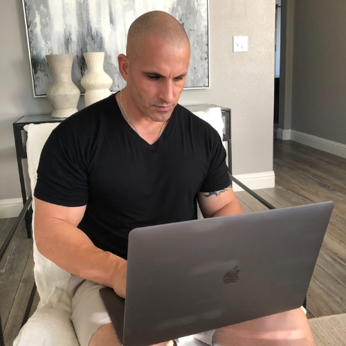 A bald man sitting on a light-colored armchair working on a grey laptop in a modern living space with light grey walls and abstract art in the background.
