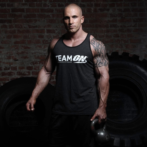 Portrait of a muscular bald man wearing a black tank top with 'TEAM ON' logo, holding a kettlebell in a dimly lit gym with tires and a brick wall.