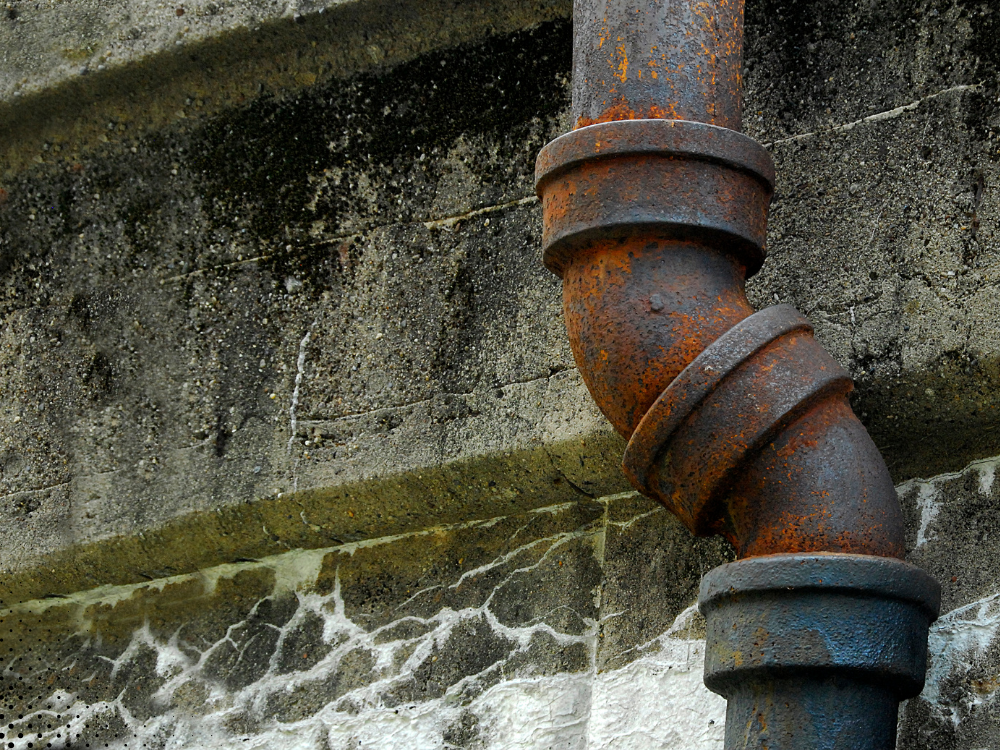 Rusty cast iron sewer pipe exterior with visible corrosion and staining on a concrete foundation wall, indicating potential drainage or plumbing issues.