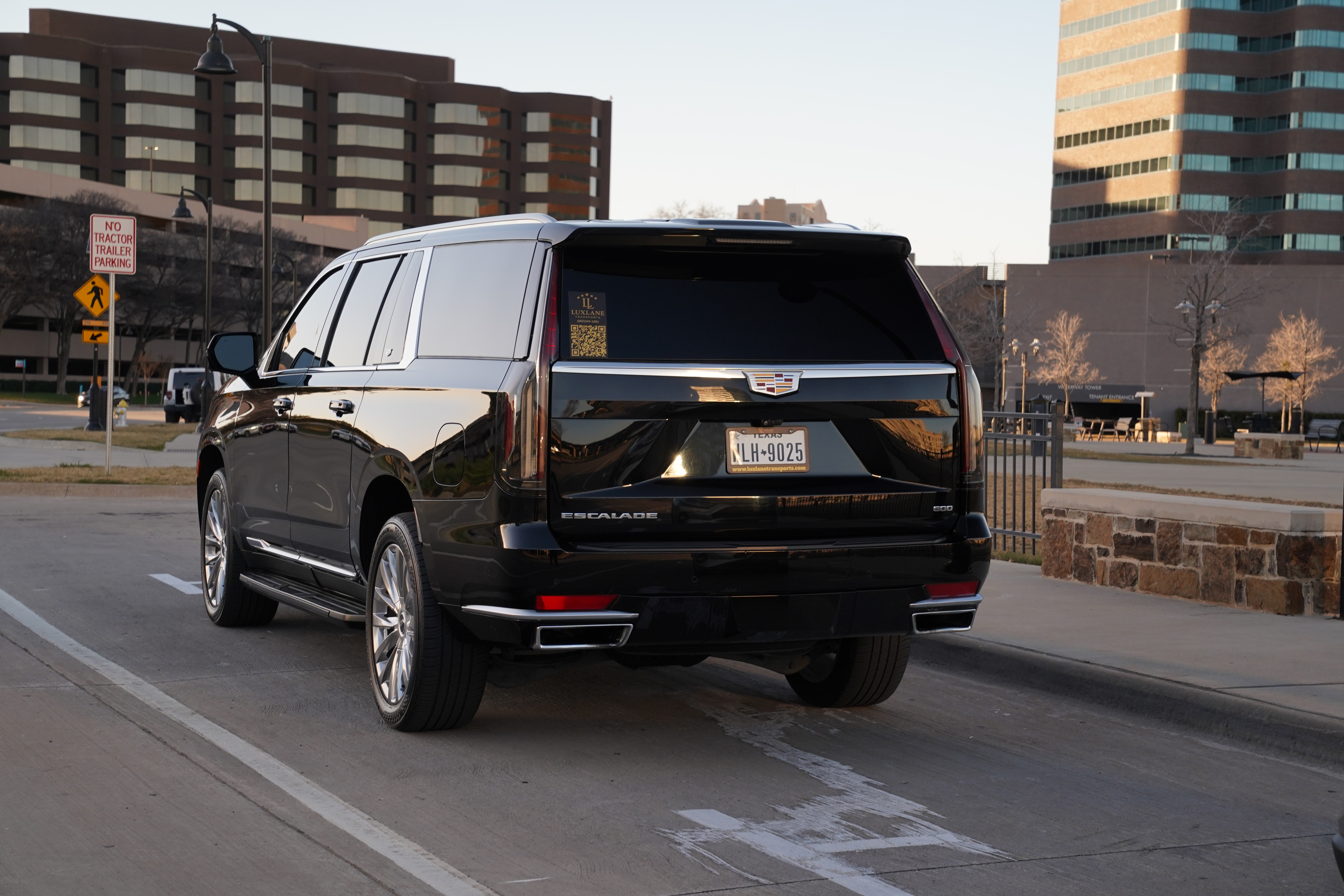 A premium full-size black GMC Yukon SUV waiting at a designated stadium pickup zone under the bright stadium arena lights.