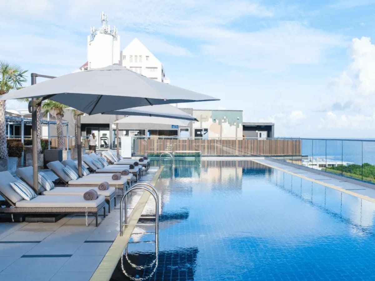 Rooftop infinity pool with lounge chairs and umbrellas, providing a stunning view of the city skyline and ocean.