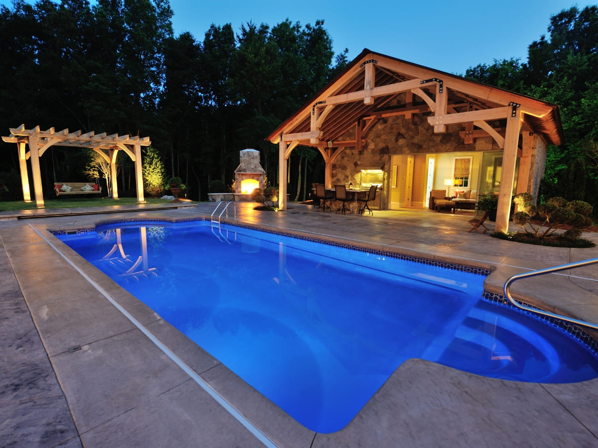 Evening view of a pool with pergola and outdoor fireplace, next to a wooden pool house in Baldwin.