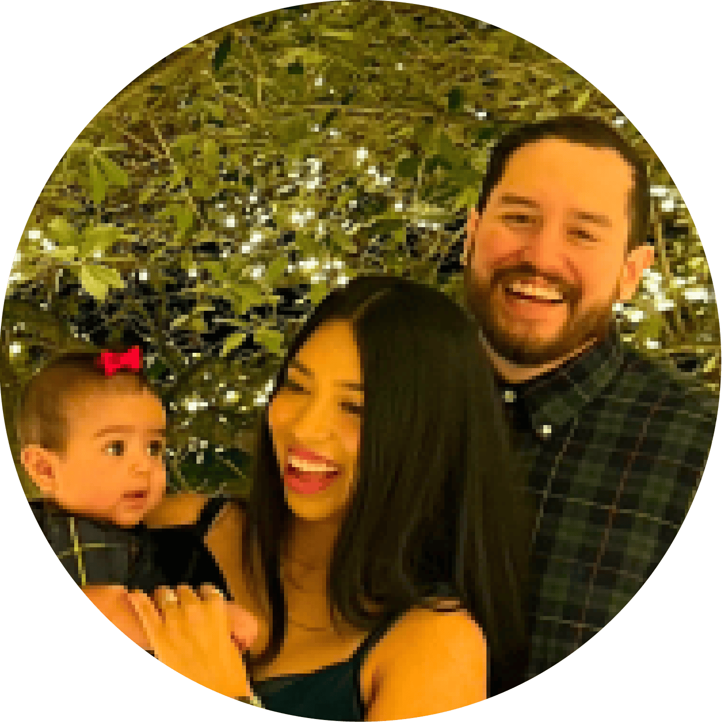 Smiling couple holding baby girl outdoors with leafy trees in background.