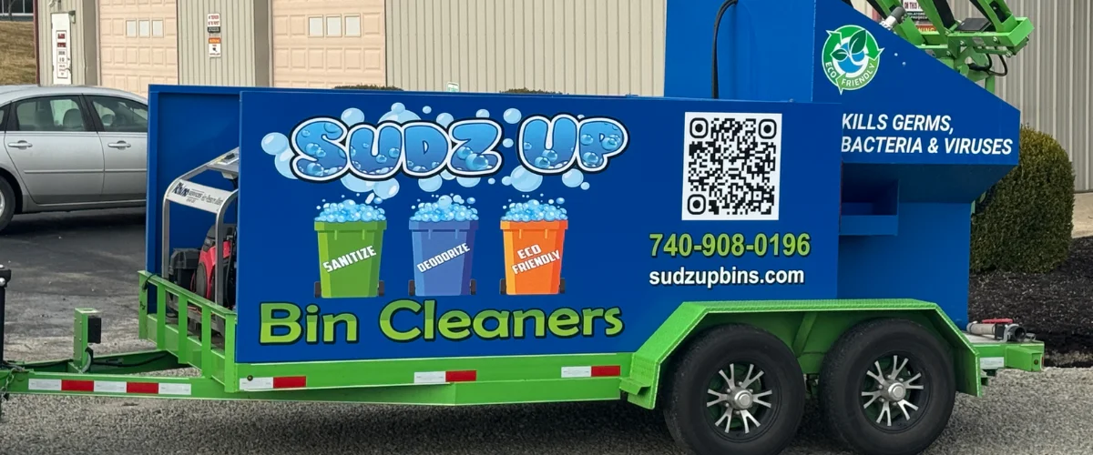 A Sudz Up Bin Cleaners trailer in blue and green, attached to a silver pickup truck, parked on gravel. A Sudz Up Bin Cleaners trailer in blue and green, attached to a silver pickup truck, parked on gravel.