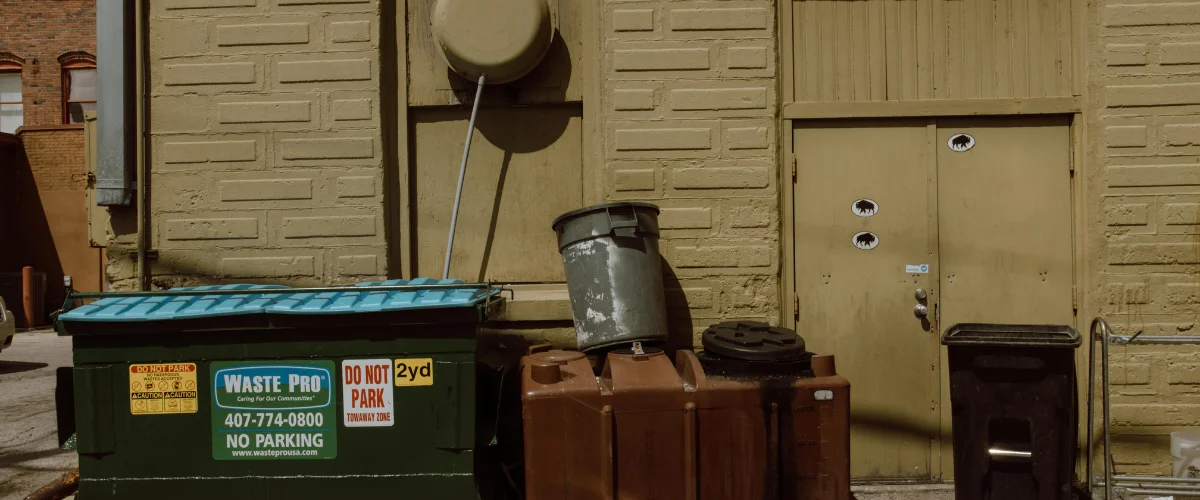 A back alley scene with two dumpsters and a brown storage bin in front of a tan brick wall and door. A back alley scene with two dumpsters and a brown storage bin in front of a tan brick wall and door.
