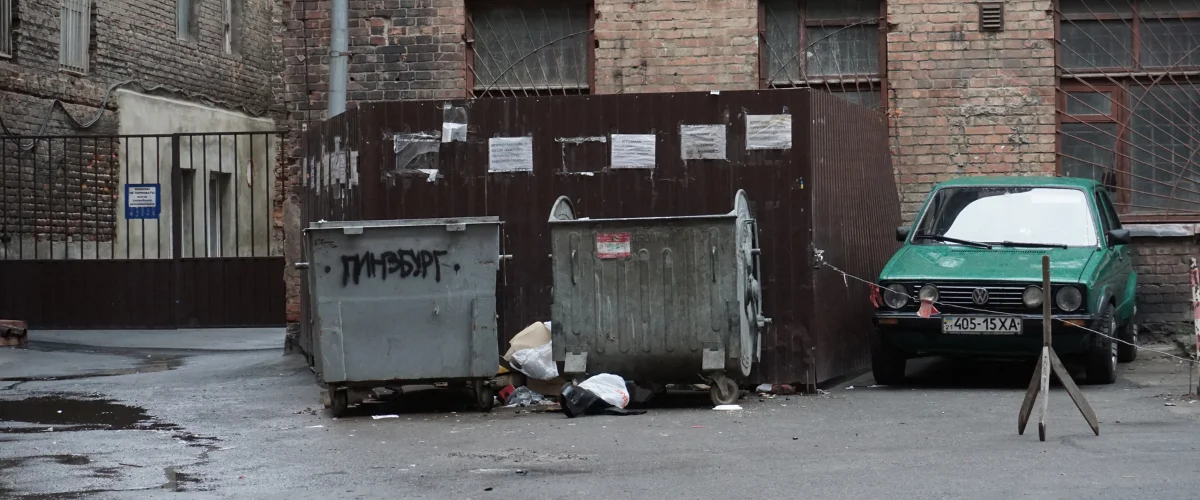 A dirty, neglected alleyway featuring two overflowing dumpsters, a run-down green car, and brick buildings. A dirty, neglected alleyway featuring two overflowing dumpsters, a run-down green car, and brick buildings.