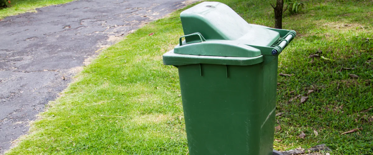 A tall, dark green plastic trash can with its lid open, sitting on a lush green lawn next to a dark asphalt path. A tall, dark green plastic trash can with its lid open, sitting on a lush green lawn next to a dark asphalt path.