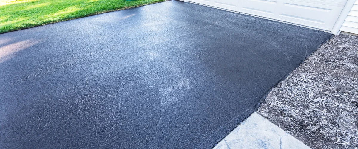 A newly seal-coated black asphalt driveway next to a green lawn and white garage door.