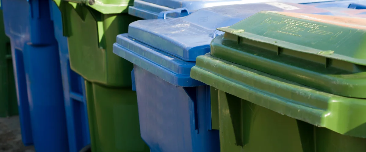 A close-up view of alternating green and blue plastic recycling bins with sunlight highlighting their lids. A close-up view of alternating green and blue plastic recycling bins with sunlight highlighting their lids.