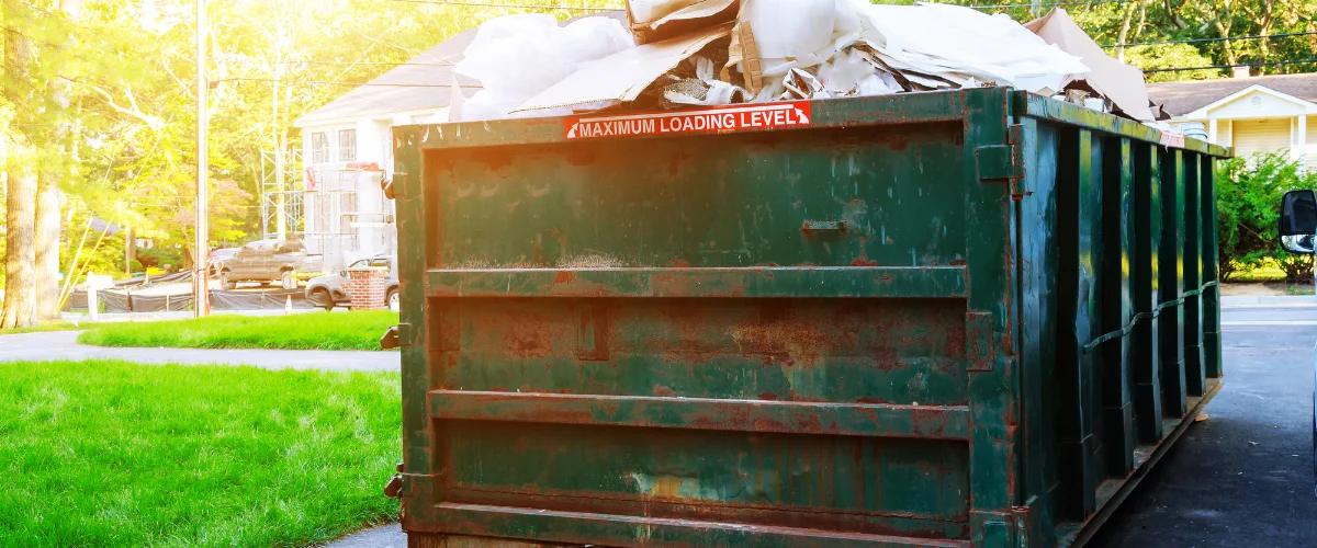 A large, dark green dumpster overflowing with construction debris and cardboard on a sunny day. A large, dark green dumpster overflowing with construction debris and cardboard on a sunny day.