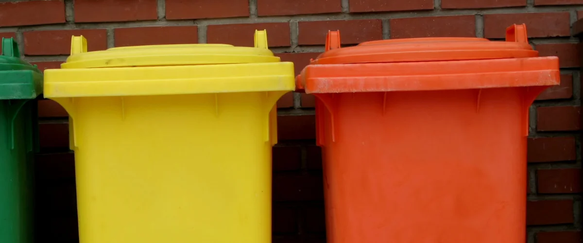 A close-up of green, yellow, and orange plastic garbage bins lined up against a red brick wall. A close-up of green, yellow, and orange plastic garbage bins lined up against a red brick wall.