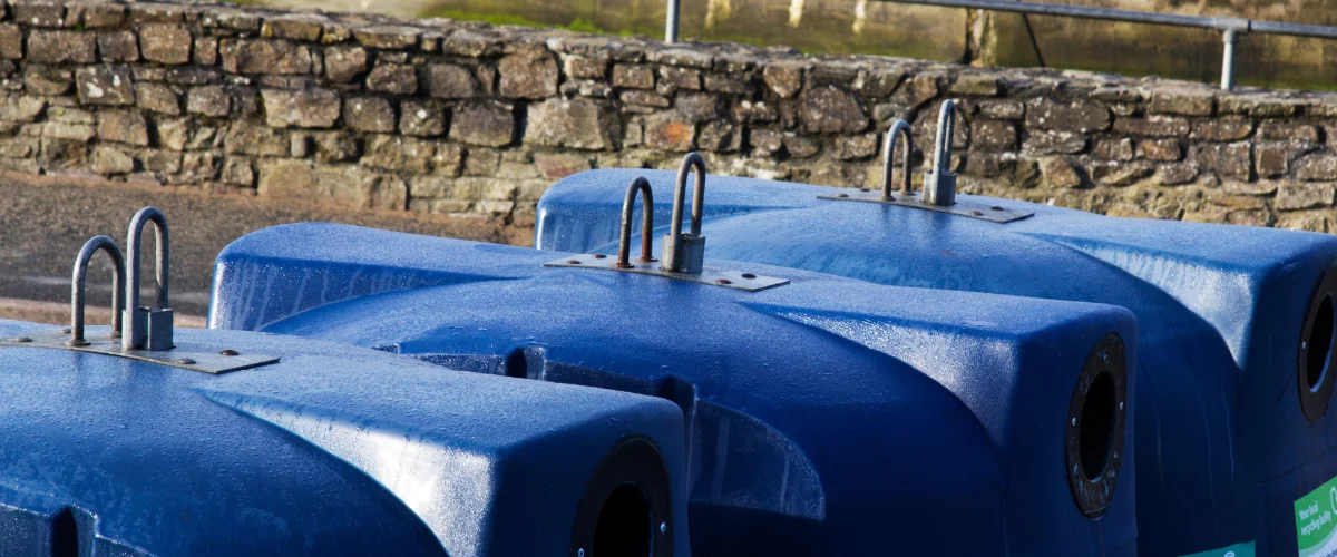 Several blue recycling bins with condensation on their surfaces sit near a stone wall. Several blue recycling bins with condensation on their surfaces sit near a stone wall.