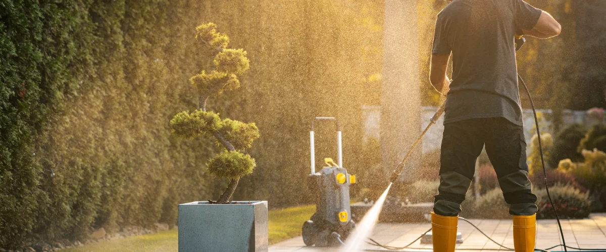 A man in gray shirt and yellow boots power-washing a patio at sunset, with a potted bonsai tree nearby. A man in gray shirt and yellow boots power-washing a patio at sunset, with a potted bonsai tree nearby.