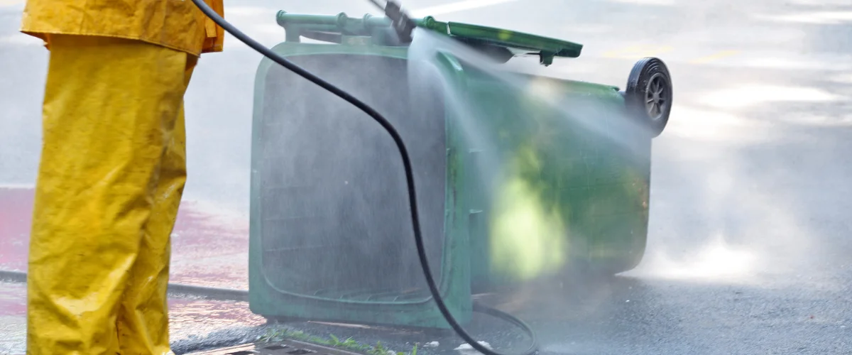 A person in yellow rain gear power washing a green garbage bin lying on its side on asphalt.