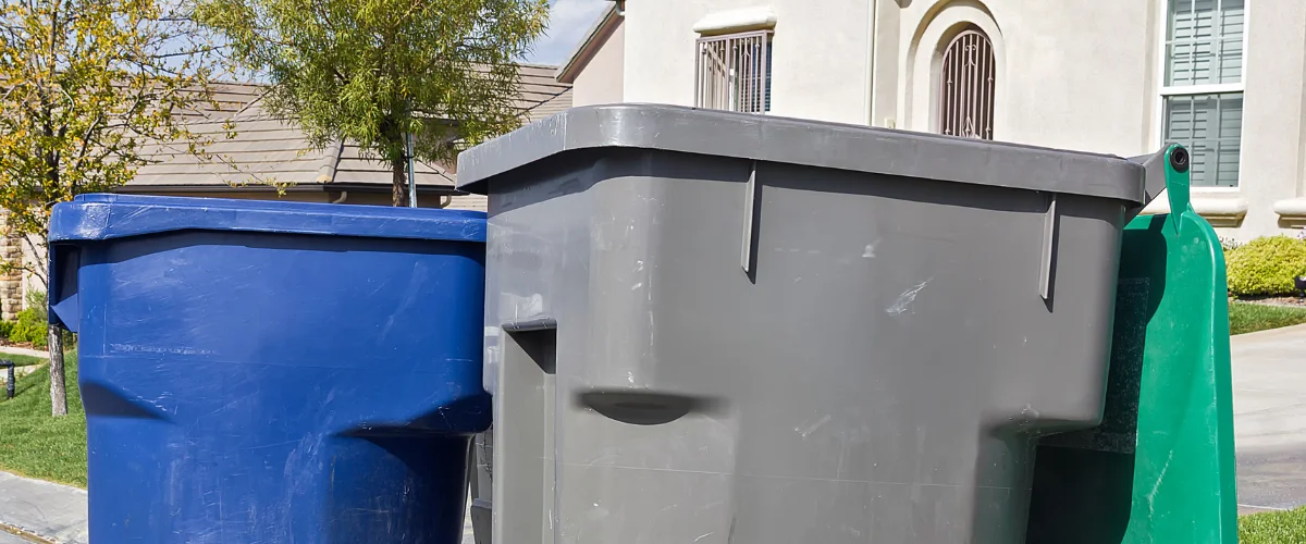 Three residential trash bins, blue, gray, and green, stand together on a sunny suburban street.