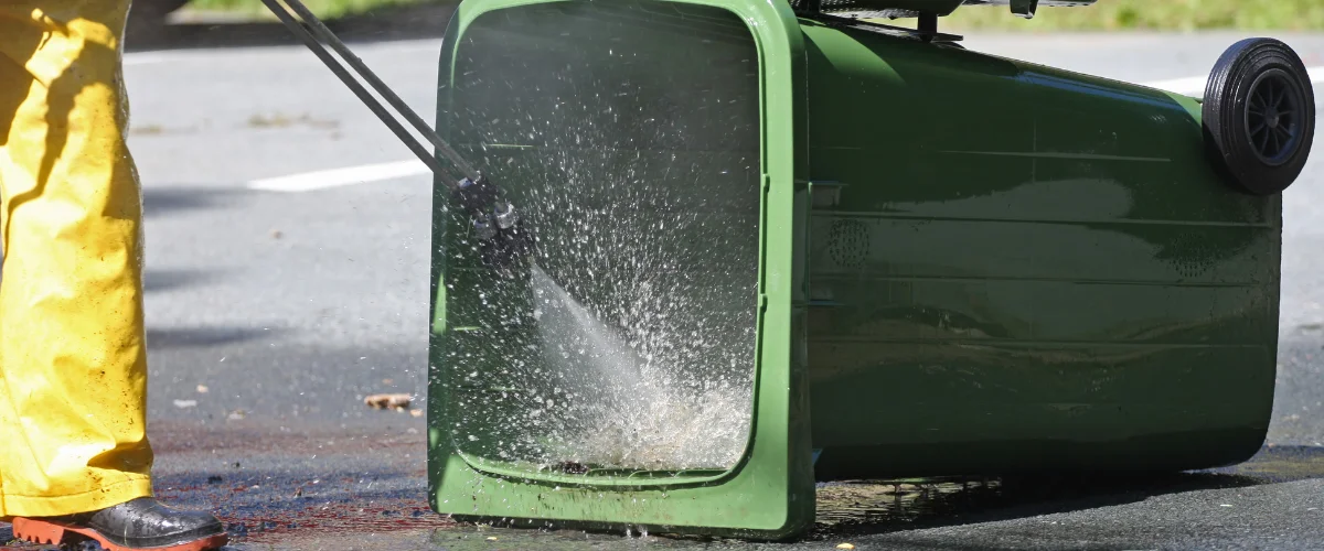 A person in yellow waders pressure washes the inside of a large green garbage bin lying on its side on asphalt.