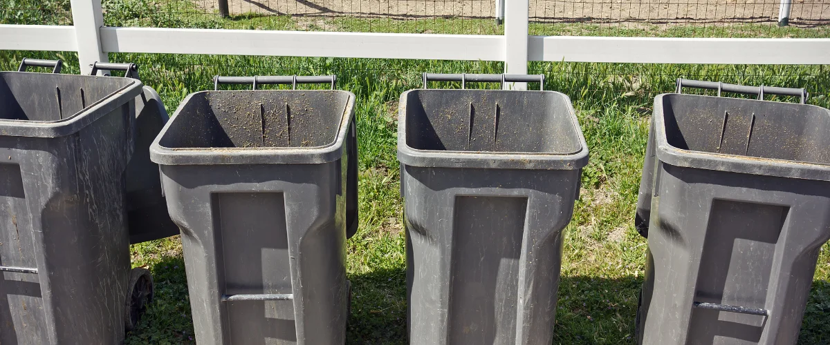 A row of four large, dirty, dark gray wheeled trash cans with open tops stands on green grass.