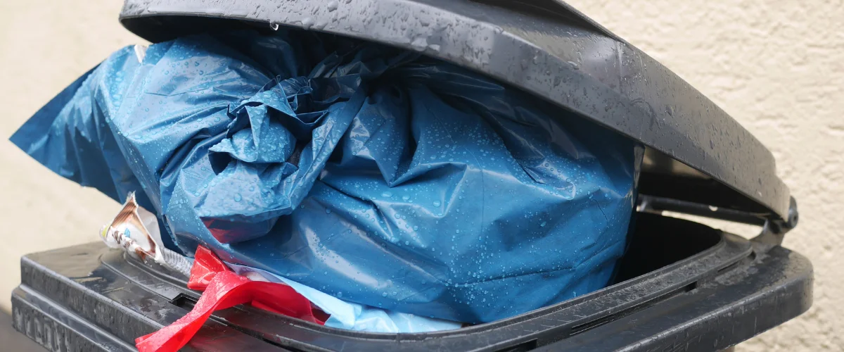 A close-up of a dark gray trash bin with an overflowing, rain-soaked blue garbage bag and a slightly ajar lid. A close-up of a dark gray trash bin with an overflowing, rain-soaked blue garbage bag and a slightly ajar lid.