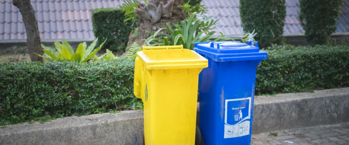 A bright yellow and a blue garbage bin with lids standing side-by-side in a lush green garden setting. A bright yellow and a blue garbage bin with lids standing side-by-side in a lush green garden setting.