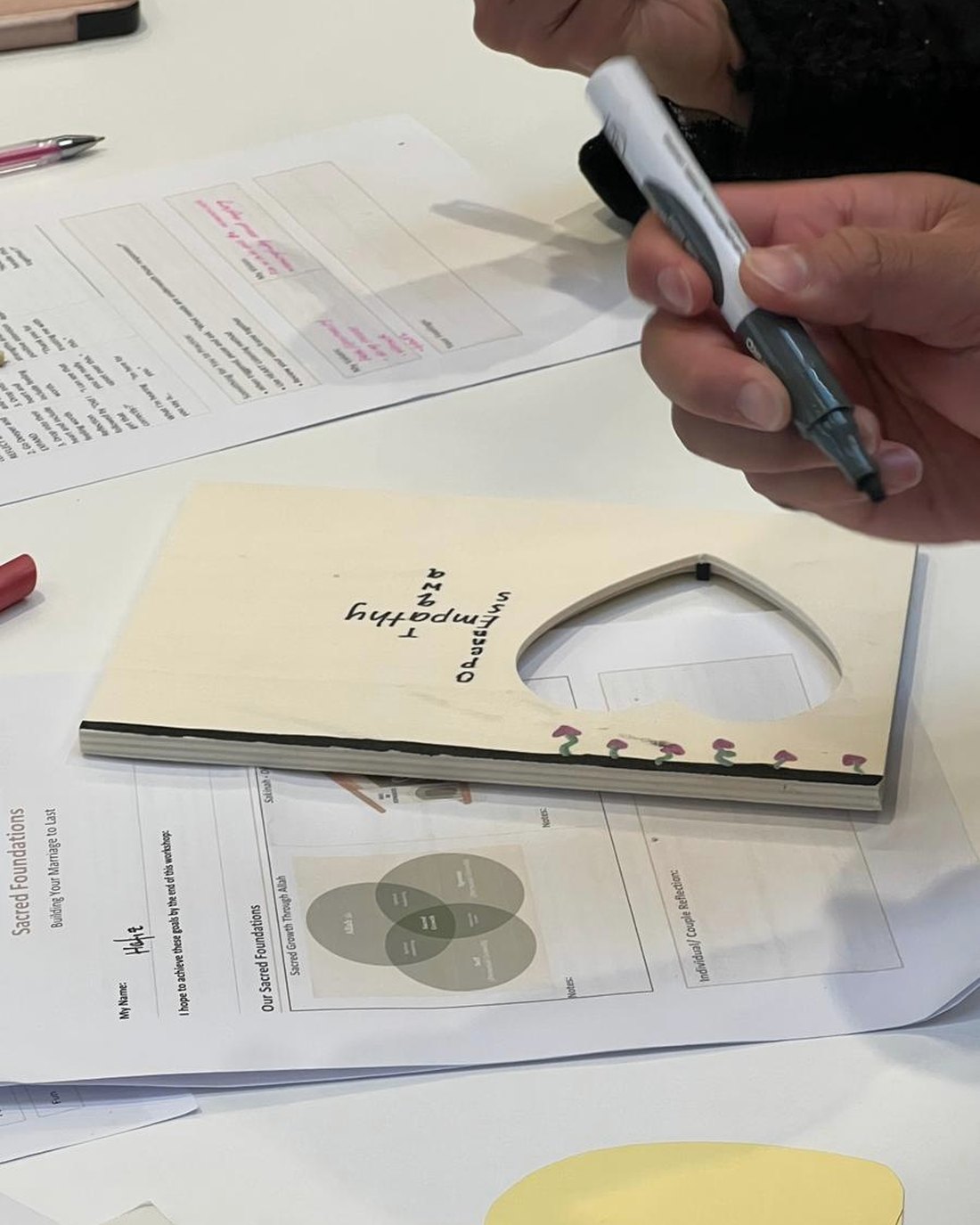 Hands at a workshop table: a marker, the Sacred Foundations worksheet and a wooden Vision Frame