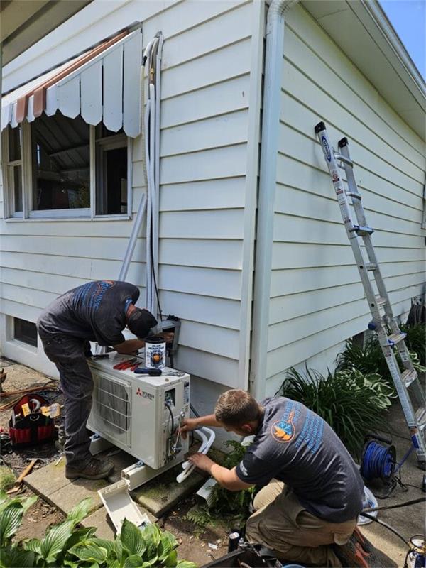 Two K.O. HVAC technicians connecting refrigerant lines on an outdoor air conditioning unit beside a white house