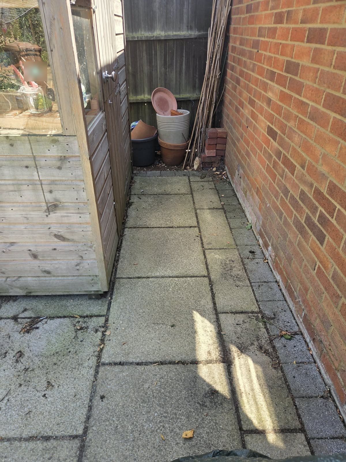 Side path of grey, grimy slabs between a timber shed and a brick wall