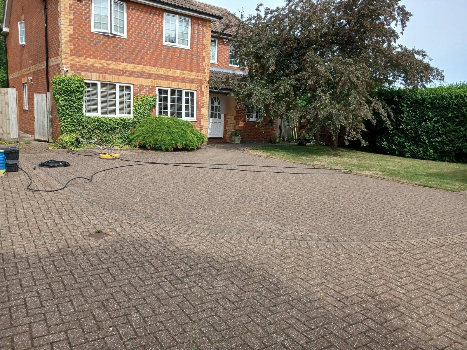 Large herringbone block-paved driveway, dark and dirty, with a hose and rotary surface cleaner laid out