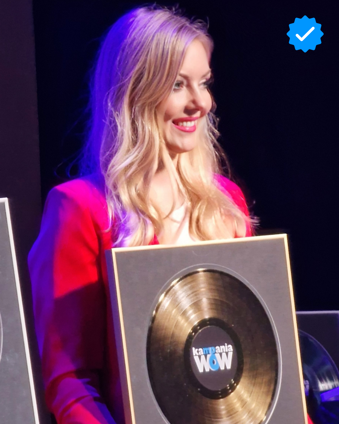 Kobieta w czerwonym garniturze uśmiecha się, trzymając w dłoniach złotą płytę z napisem „Kampania WOW” podczas uroczystej gali lub wydarzenia branżowego.