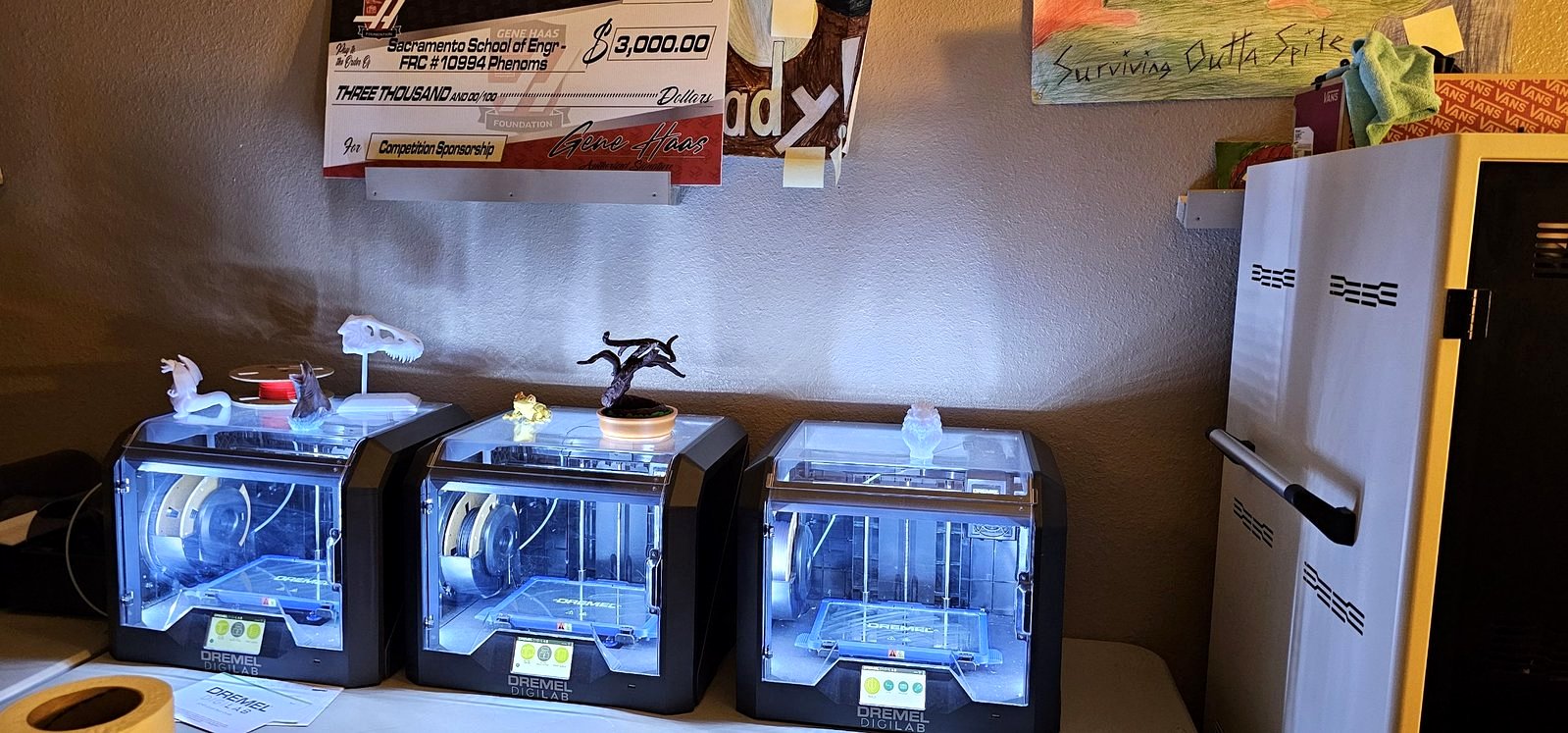 Three Dremel 3D printers with student prints on top, under a Gene Haas Foundation check for the SSES Phenoms robotics team