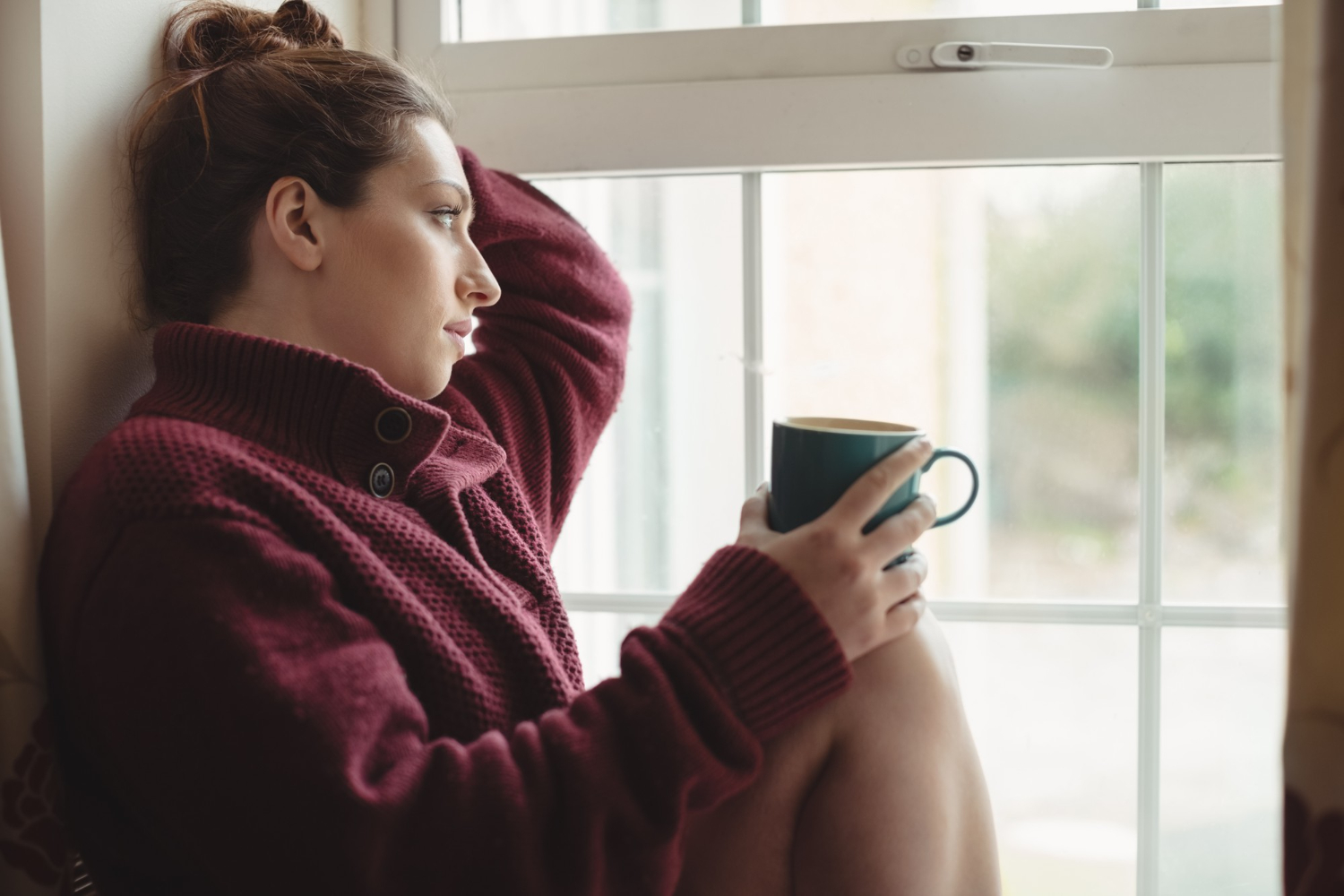 Thoughtful woman sitting at window sill and holding coffee cup Thoughtful woman sitting at window sill and holding coffee cup
