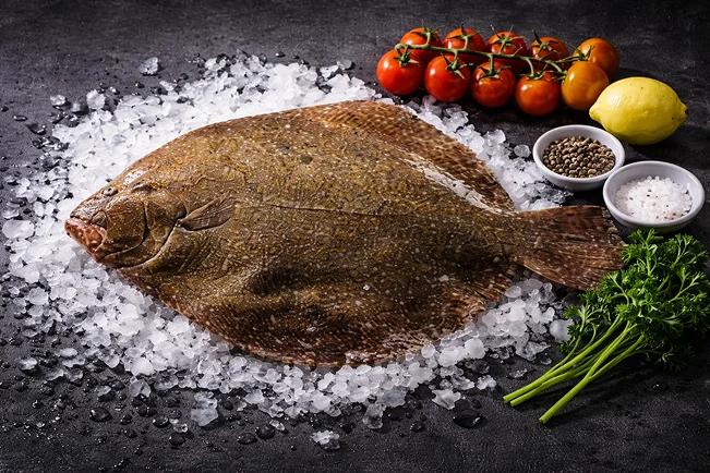 Un turbot entier et frais sur un lit de glace pilée, entouré de tomates, de citron, de persil et d’épices.